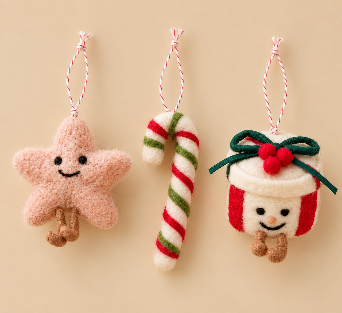 Set of 3 felted wool Christmas tree decorations: a smiley pink star, a red, white and green candy cane and a smiley present with a green bow, on a beige background